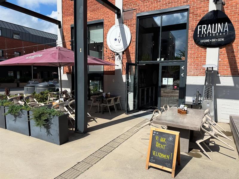 Uteservering foran restauranten med bord, stoler og blomsterkasser. Et skilt med restaurantens navn henger over inngangen, og et gatestativ viser dagens meny