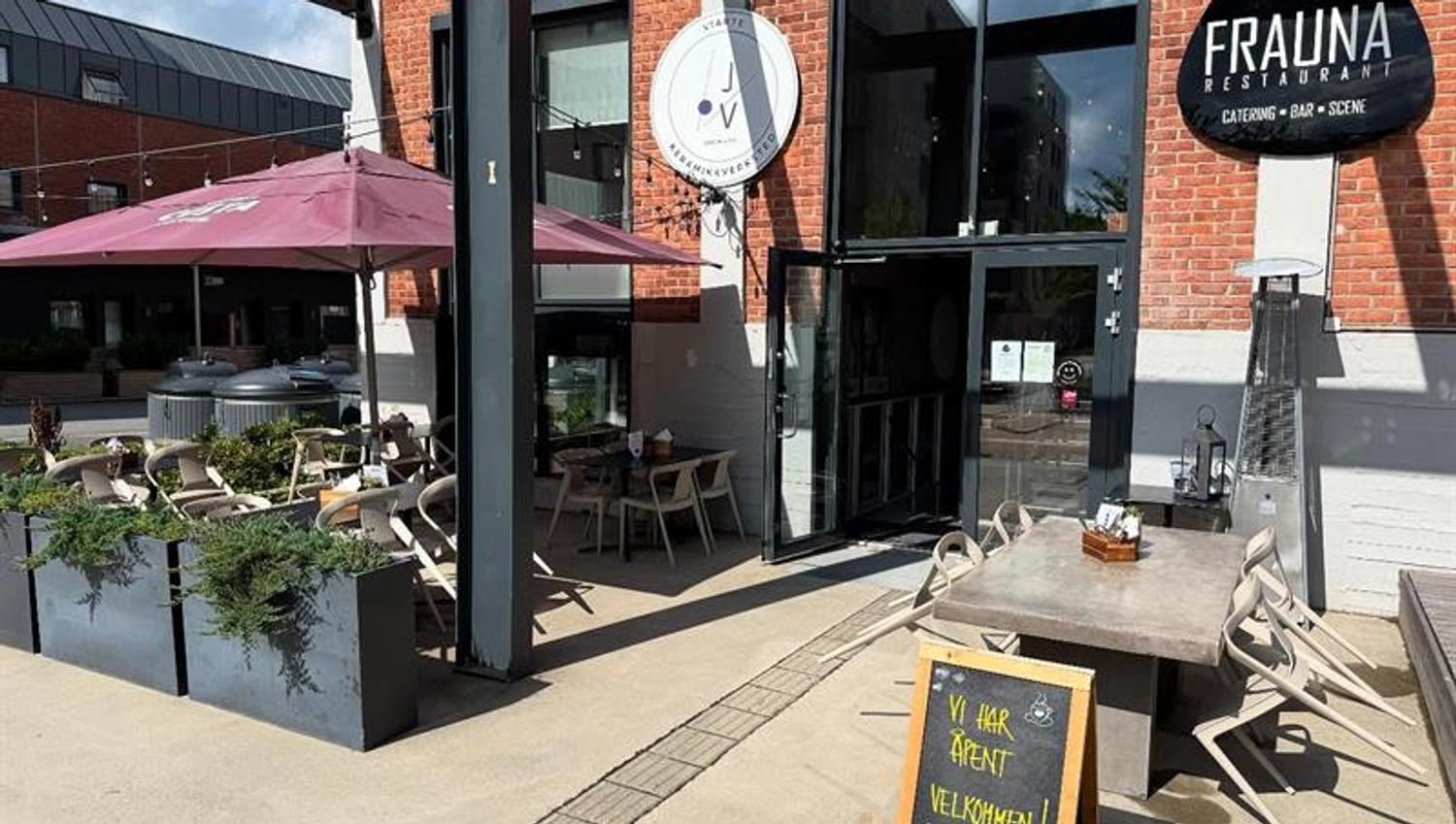Uteservering foran restauranten med bord, stoler og blomsterkasser. Et skilt med restaurantens navn henger over inngangen, og et gatestativ viser dagens meny