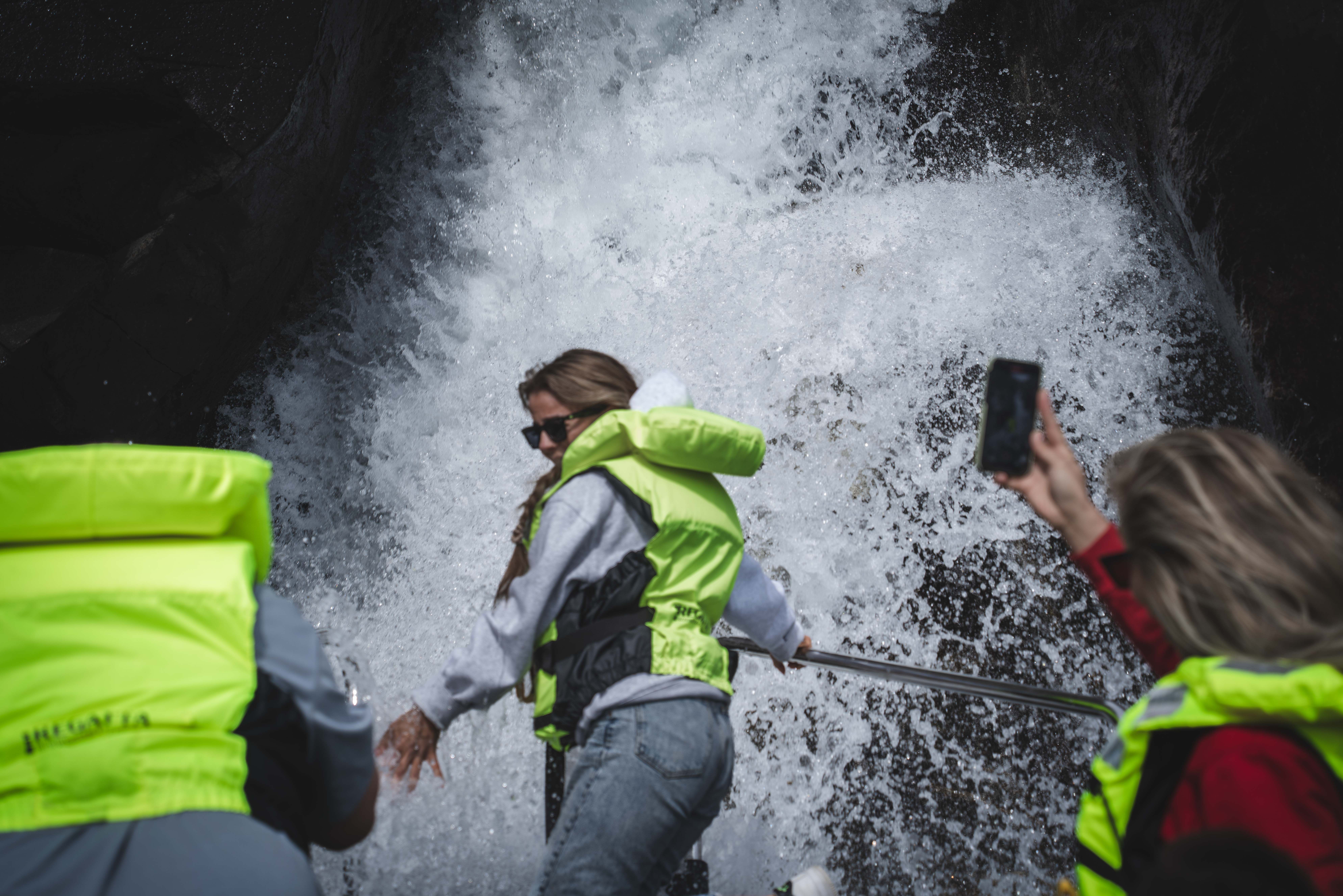 En kvinne foran i båten blir «dusjet» av en foss, andre passasjerer tar bilder med telefonene sine.
