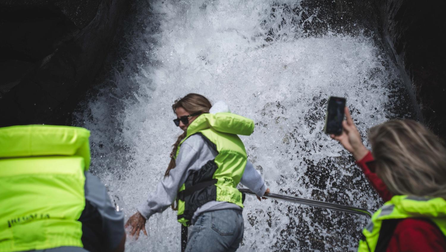 En kvinne foran i båten blir «dusjet» av en foss, andre passasjerer tar bilder med telefonene sine.