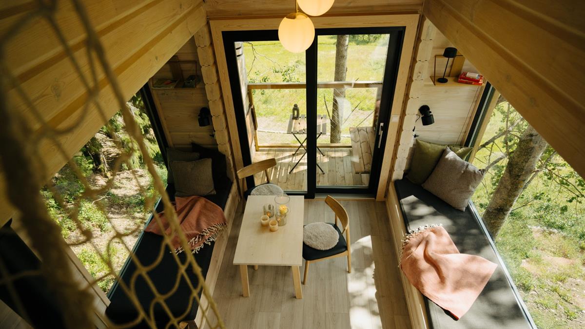 Bright cabin interior with a dining table and seating area, viewed from above. Large windows provide a view of the forest and nature.
