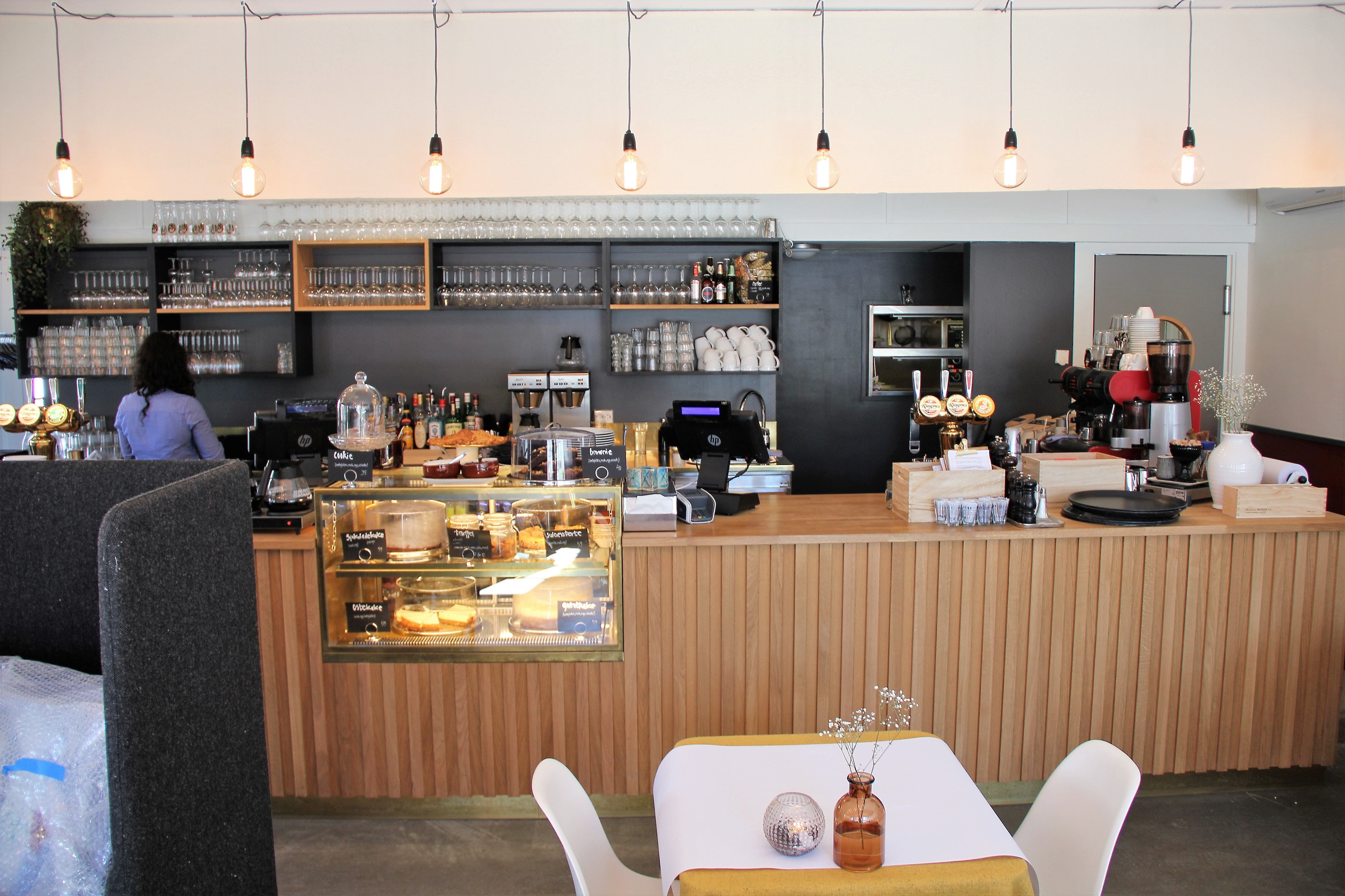 Interiør av en kafé med bar, glassmonter med kaker, kaffemaskin, glasshyller og hengende lyspærer..