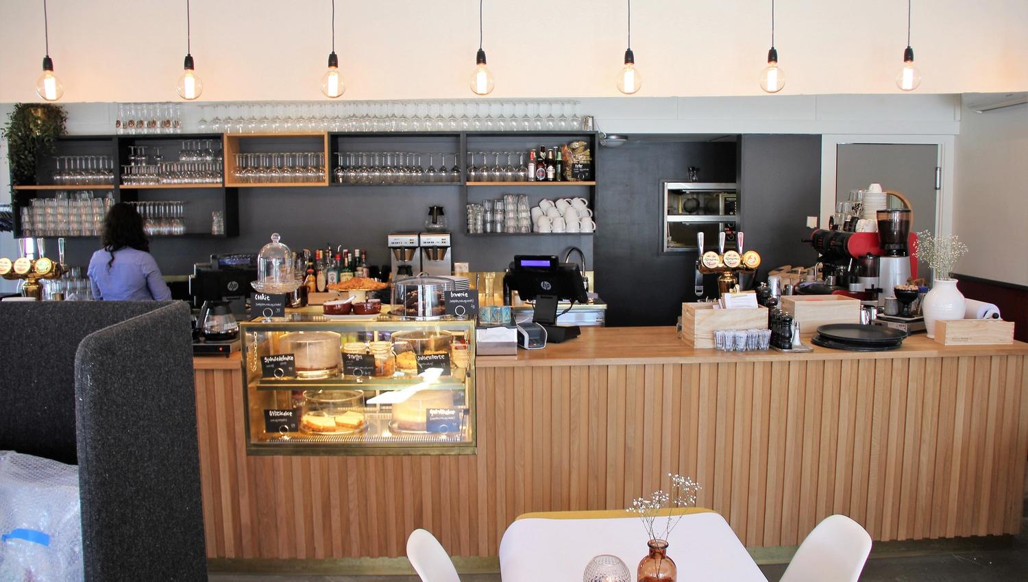 Interiør av en kafé med bar, glassmonter med kaker, kaffemaskin, glasshyller og hengende lyspærer..