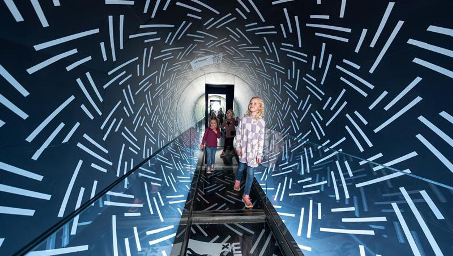Barn går gjennom en speilbelagt tunnel med lysmønstre som gir en futuristisk og sanselig opplevelse.