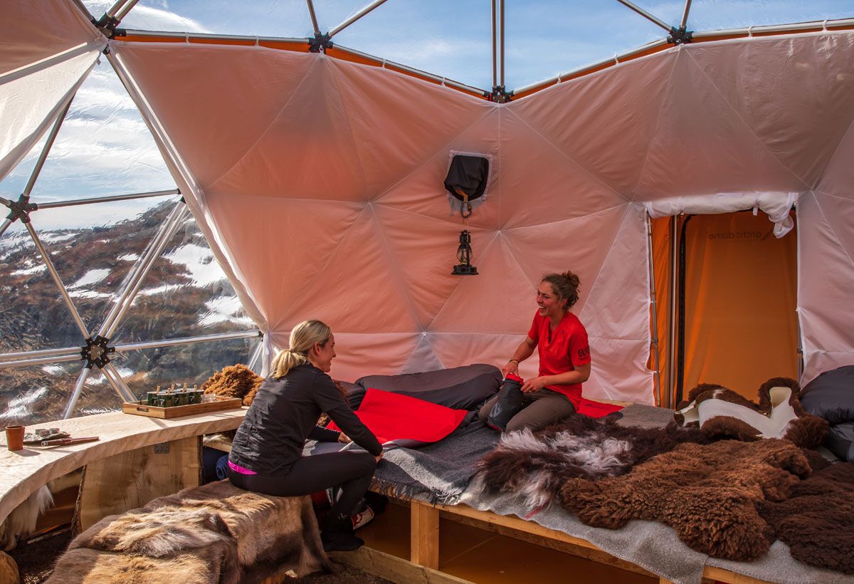 Two women chatting leisurely in a comfortable dome tent overlooking snow-capped mountains, with furs on the seating.