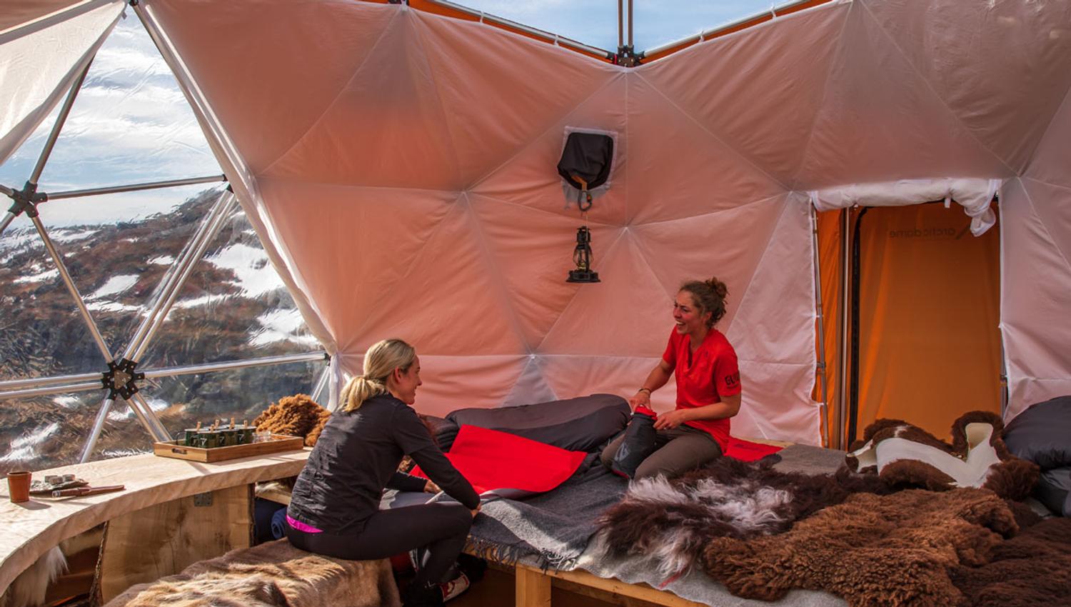 Two women chatting leisurely in a comfortable dome tent overlooking snow-capped mountains, with furs on the seating.