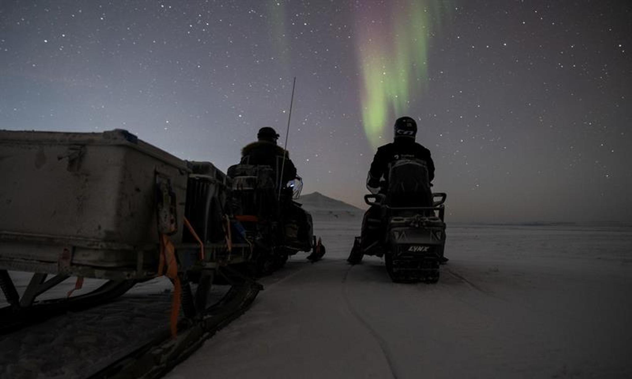 Two people sitting on snowmobiles and looking at the northern lights on the sky