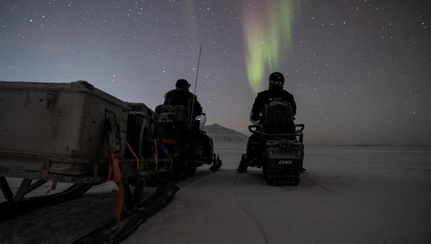 Two people sitting on snowmobiles and looking at the northern lights on the sky
