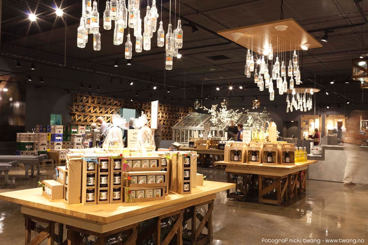 Et moderne utsalgssted med store trebord fulle av lokale produkter som syltetøy, saft og flasker med drikke. Over bordene henger lamper laget av glassflasker, og gulvet skinner i lyset..