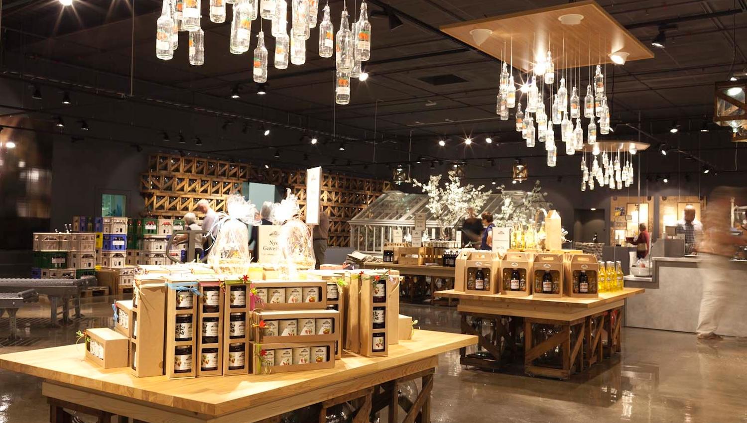 Et moderne utsalgssted med store trebord fulle av lokale produkter som syltetøy, saft og flasker med drikke. Over bordene henger lamper laget av glassflasker, og gulvet skinner i lyset..