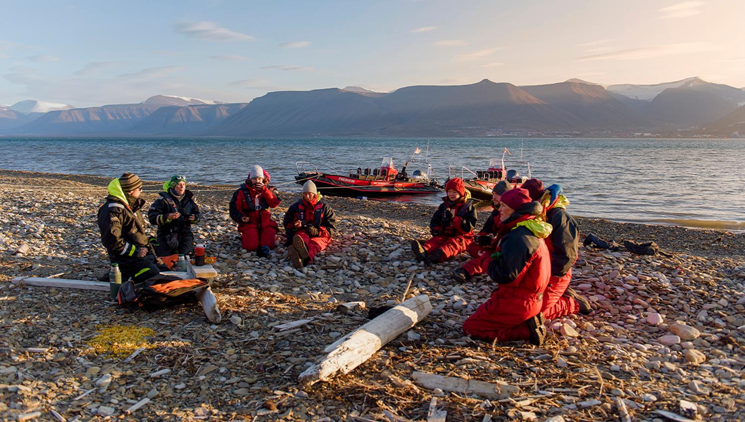 A group of guests and two guides sitting in a circle on a beach talking to each other, with two RIB boats anchored by the shore and a fjord in the bac