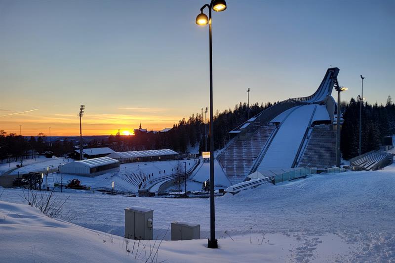 Sunset over the Holmenkollen ski jump on a cold winter evening, with snow-covered stands and lights glowing at dusk.