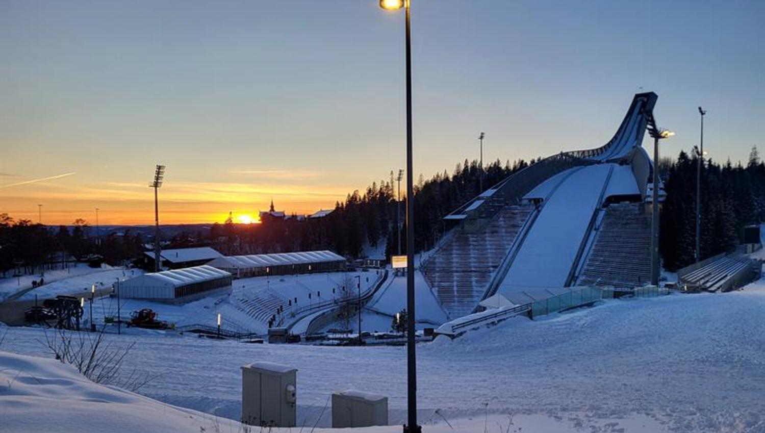 Sunset over the Holmenkollen ski jump on a cold winter evening, with snow-covered stands and lights glowing at dusk.