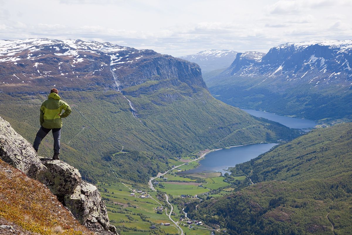 A man stands on a rocky outcrop in a steep hillside and enjoyes a magnificent view into a steep valley with a lake on the bottom.