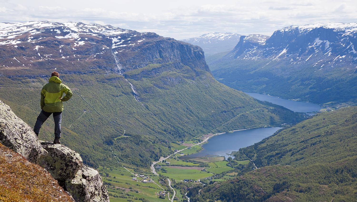 A man stands on a rocky outcrop in a steep hillside and enjoyes a magnificent view into a steep valley with a lake on the bottom.