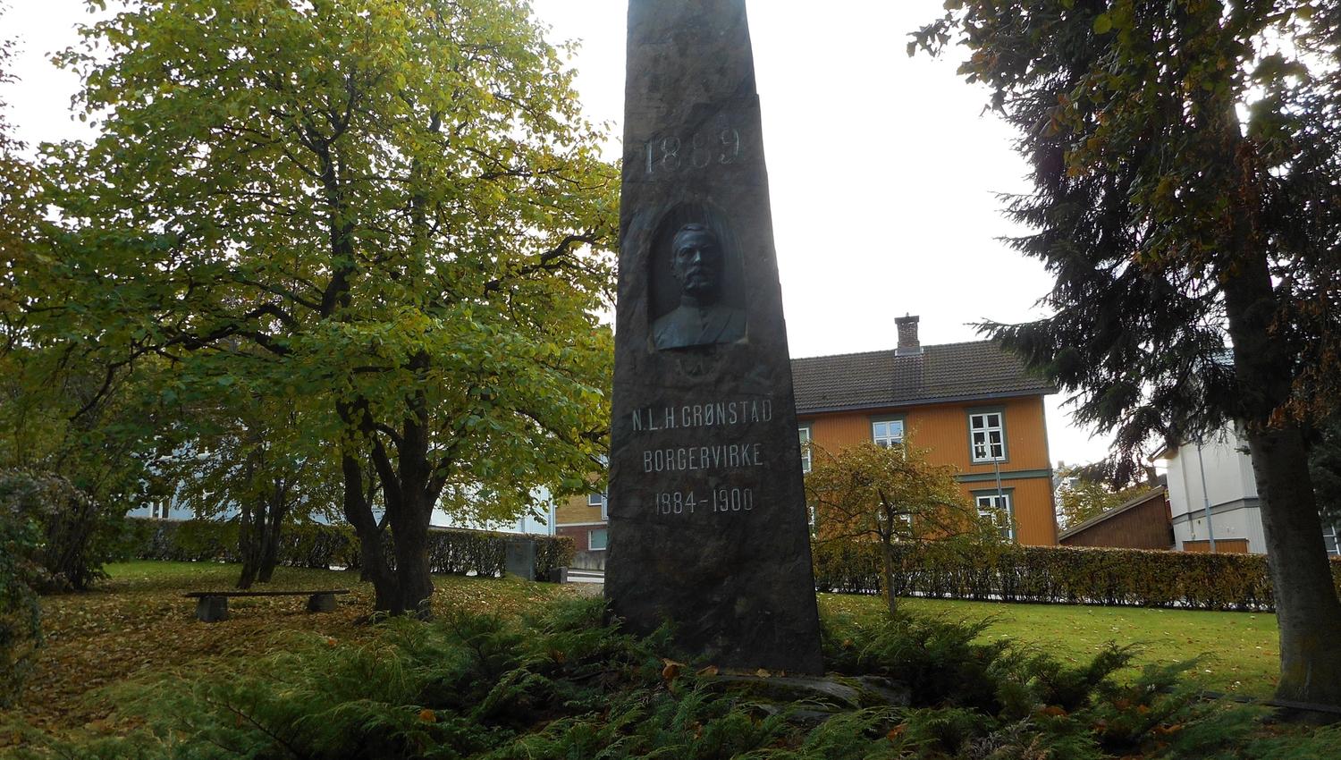 Høy, mørk minnestein med innskrift i en gresspark med trær og bygninger i bakgrunnen på høsten..