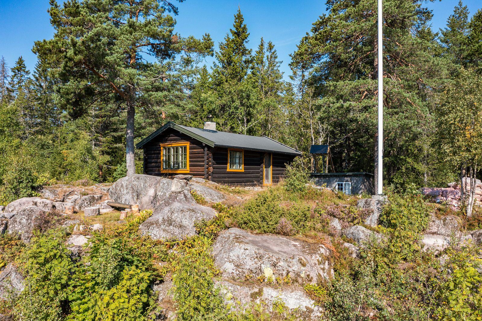 Brun markahytte og flaggstang på fjellknaus omgitt av grønn vegetasjon