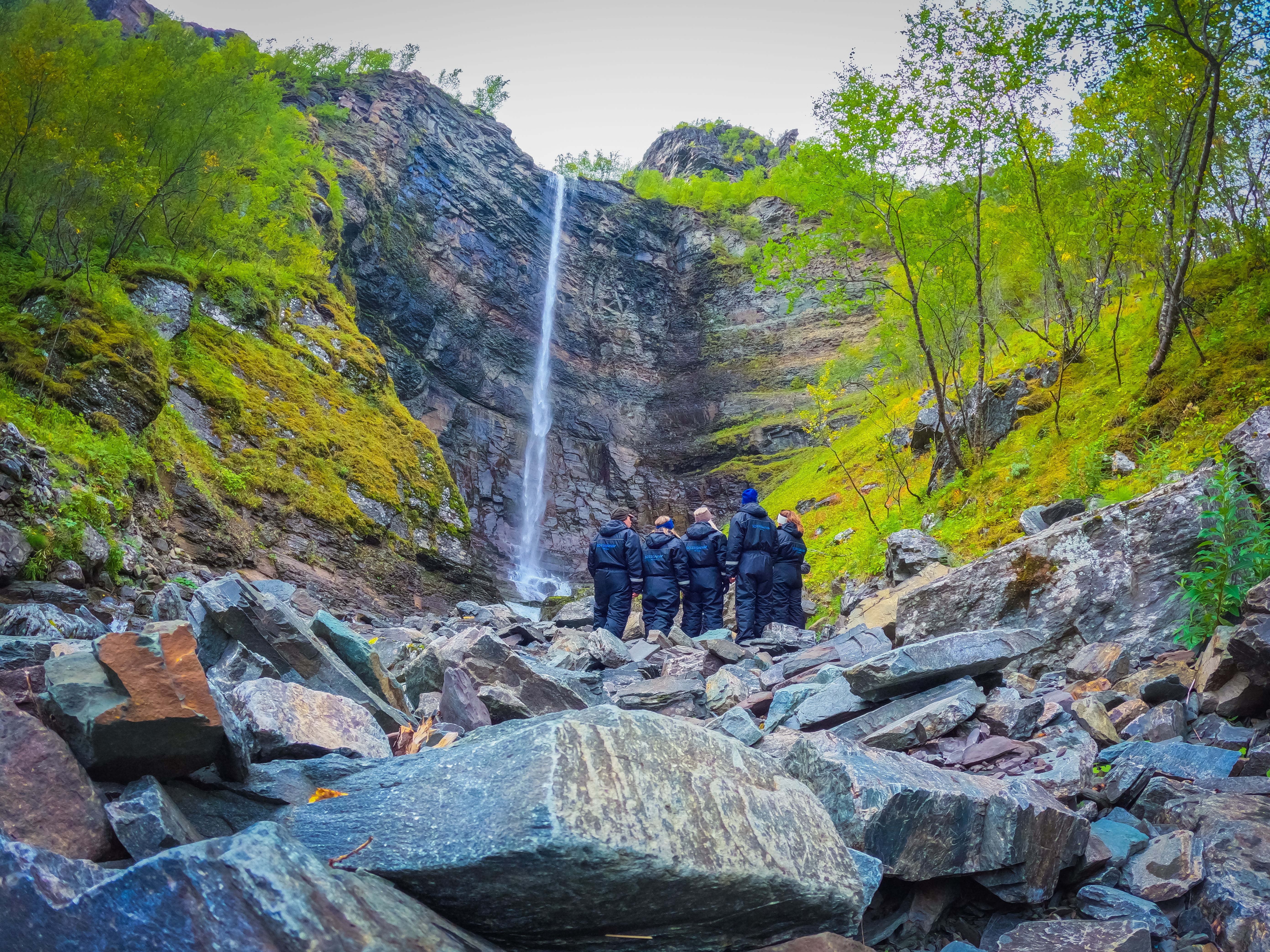 Experience the tranquility of nature as a stunning waterfall flows down majestic cliffs. Adventurers clad in jackets explore the rocky terrain, surrounded by vibrant foliage and the sounds of cascading water. A perfect spot for those seeking adventure and
