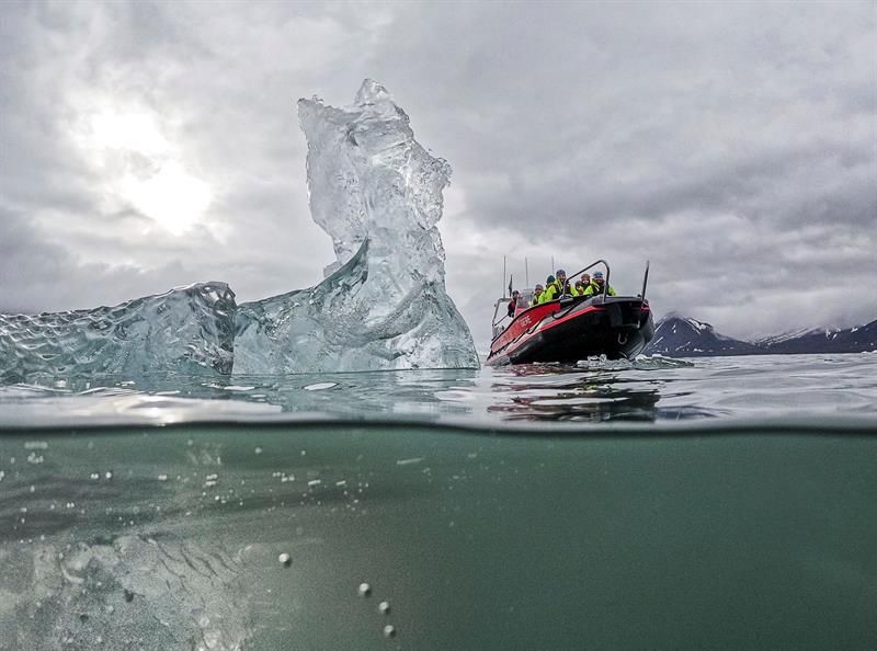 Halv-undersjøisk bilde av isfjell med RIB-båt ved siden av i gråvær.
