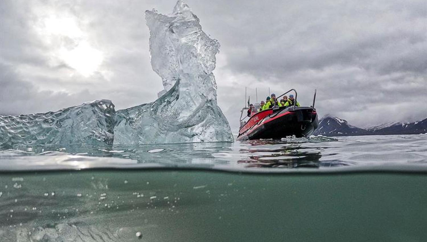 Halv-undersjøisk bilde av isfjell med RIB-båt ved siden av i gråvær.