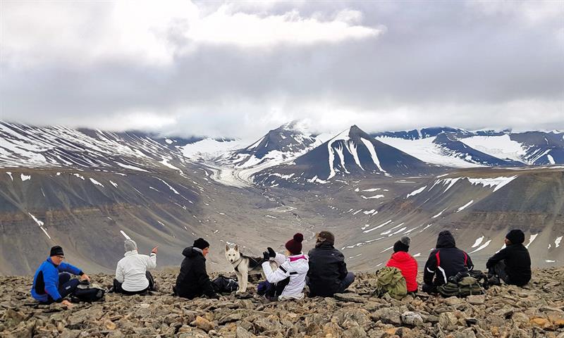Åtte mennesker og en hund som sitter på kanten av et fjell, med en dyp dal og fjell med litt snø i bakgrunnen