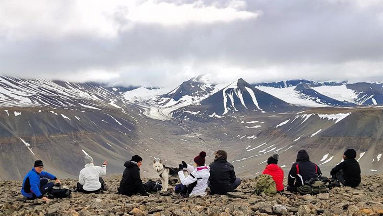 Åtte mennesker og en hund som sitter på kanten av et fjell, med en dyp dal og fjell med litt snø i bakgrunnen