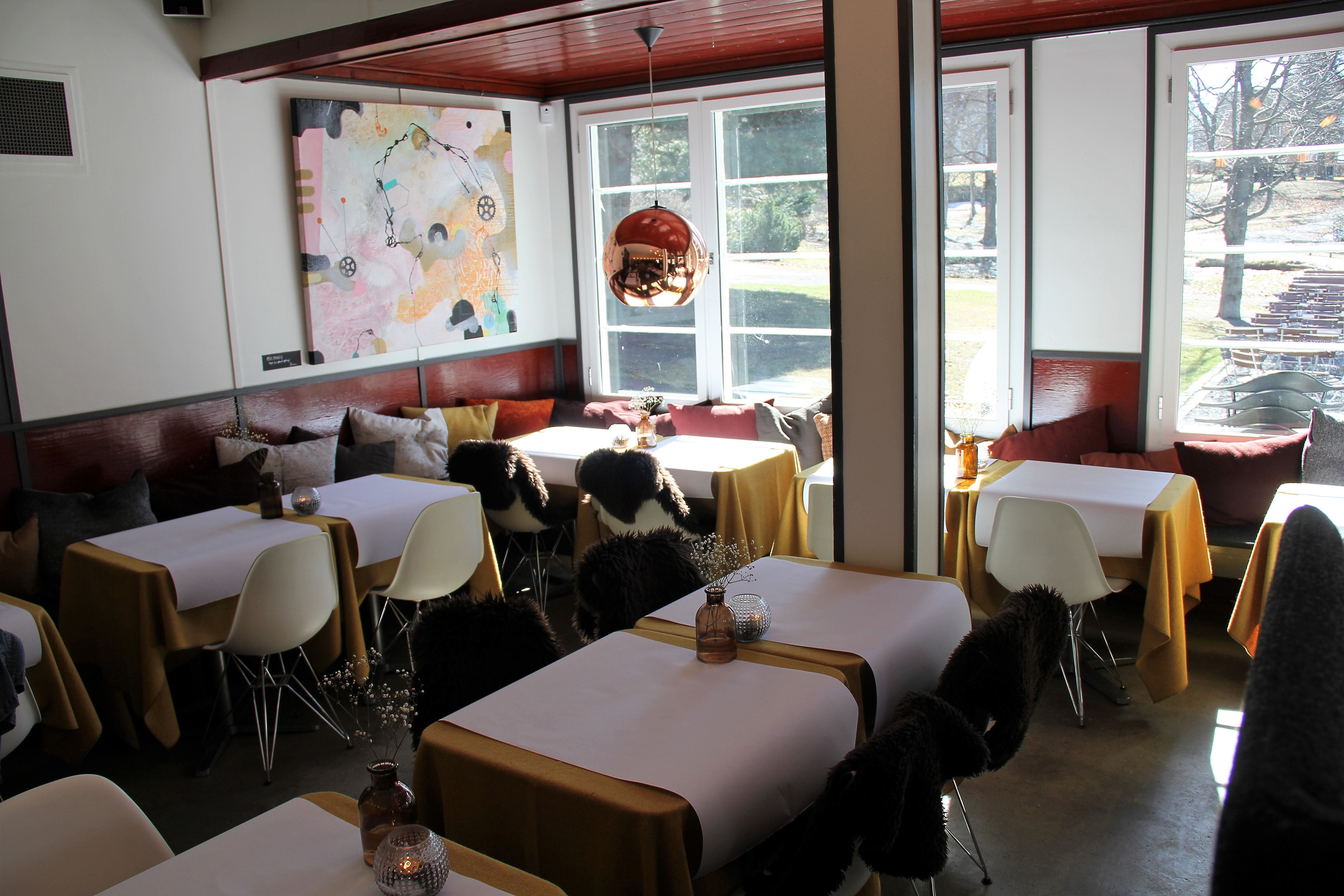 Lyst kafé- eller restaurantinteriør med borddukede bord, hvite stoler, puter langs veggen og store vinduer..