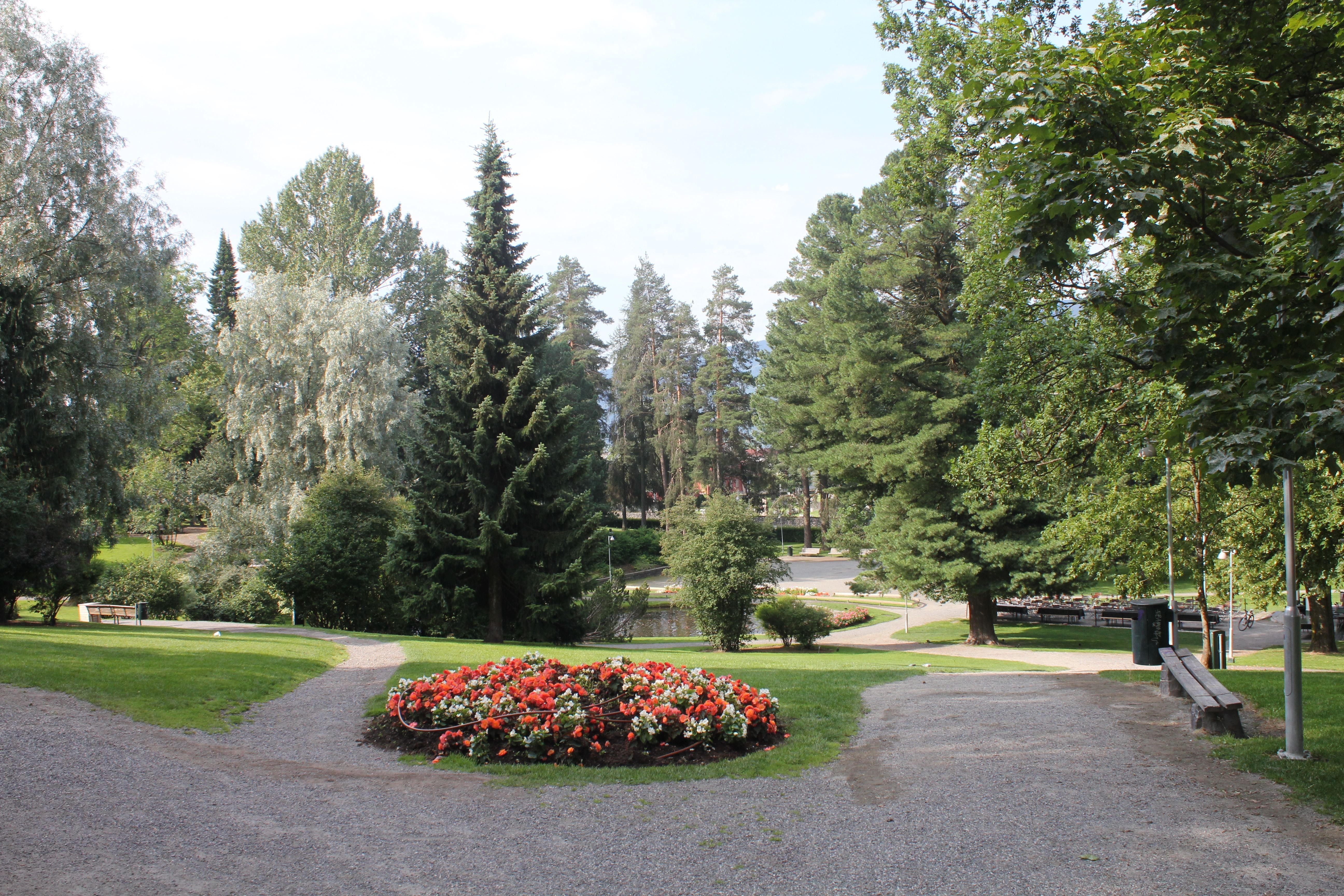 Grussti i en grønn park med et rundt blomsterbed i røde og hvite blomster, omgitt av høye trær..