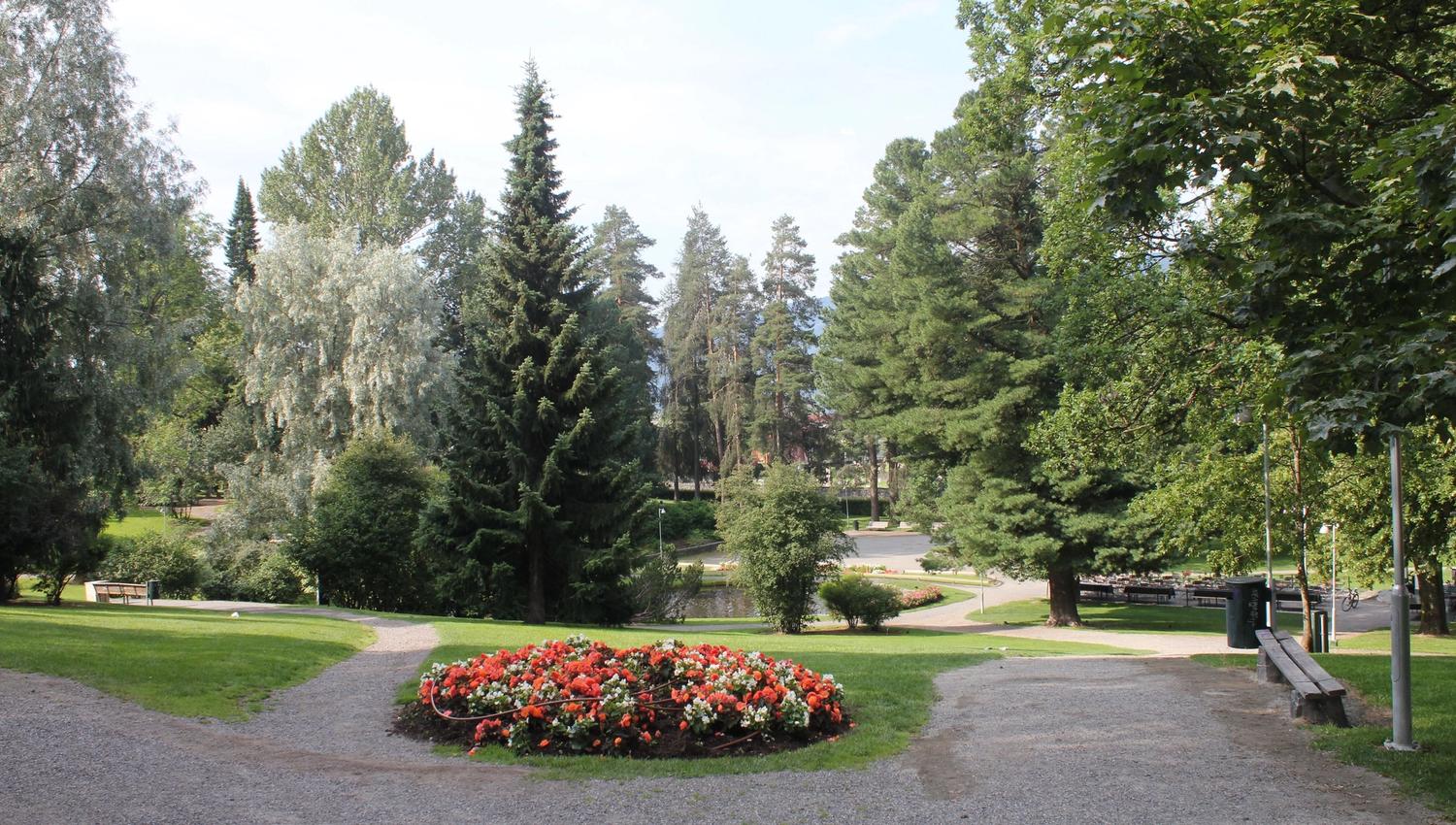 Grussti i en grønn park med et rundt blomsterbed i røde og hvite blomster, omgitt av høye trær..