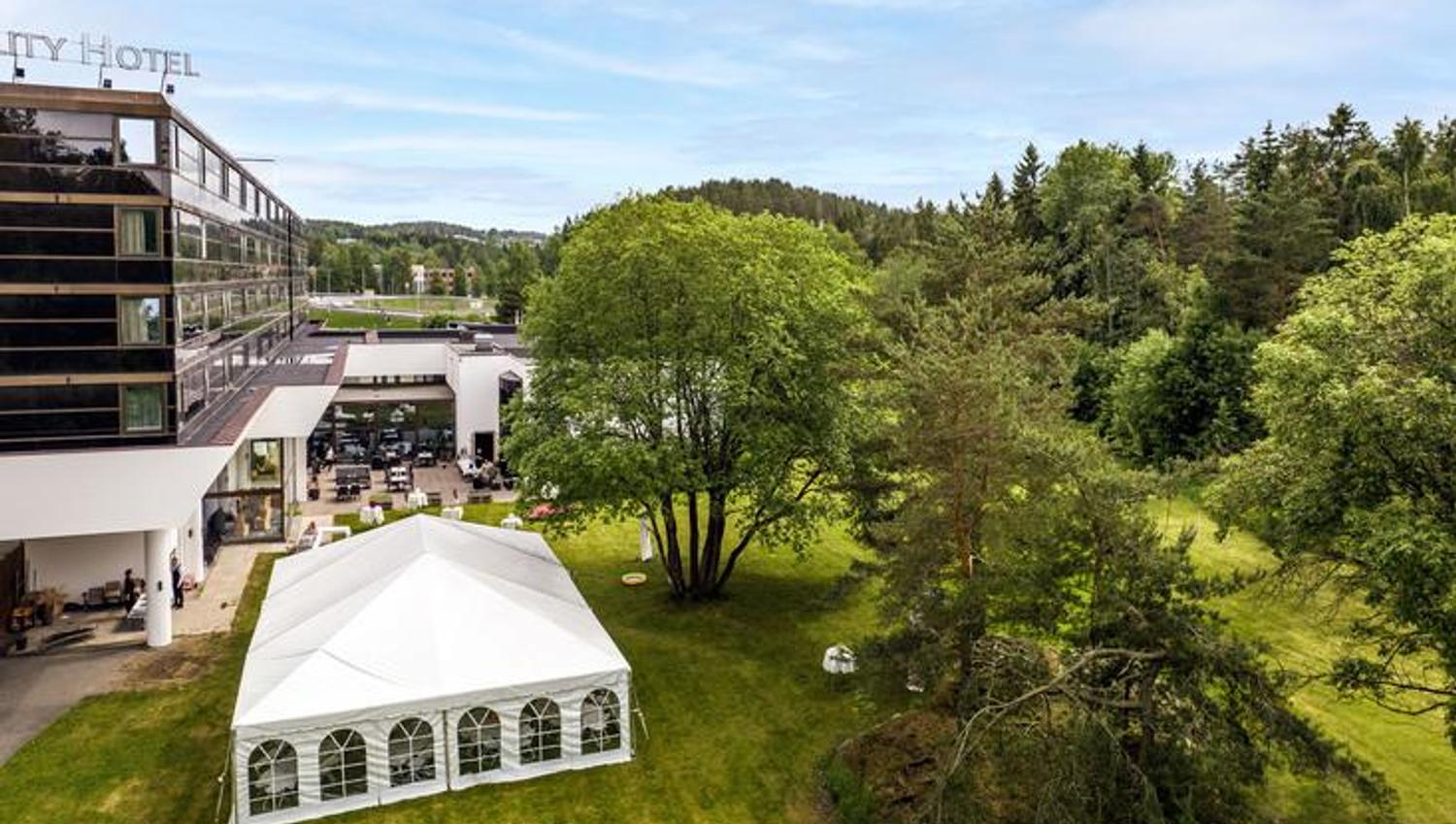Grønt uteområde med trær og et hvitt telt, sett ovenfra i naturskjønne omgivelser.