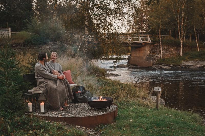 To mennesker sitter på en benk ved elva på Ådalsvollen Retreat, ikledd morgenkåper, og snakker sammen ved bålpanna og tente stearinlys med en liten bro og trær i bakgrunnen.