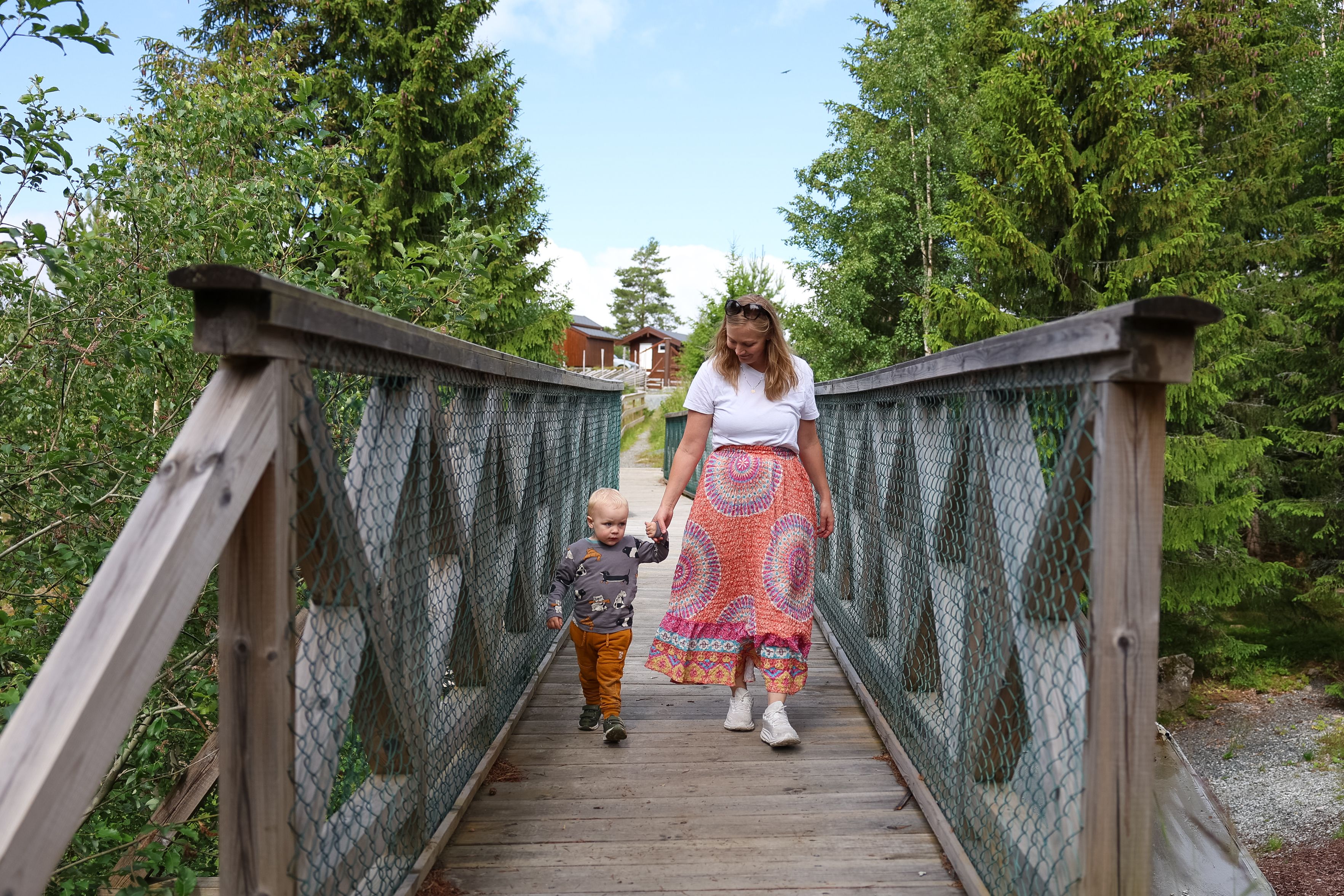 Child and an adult walking across a wooden bridge surrounded by green forest in the nature park.