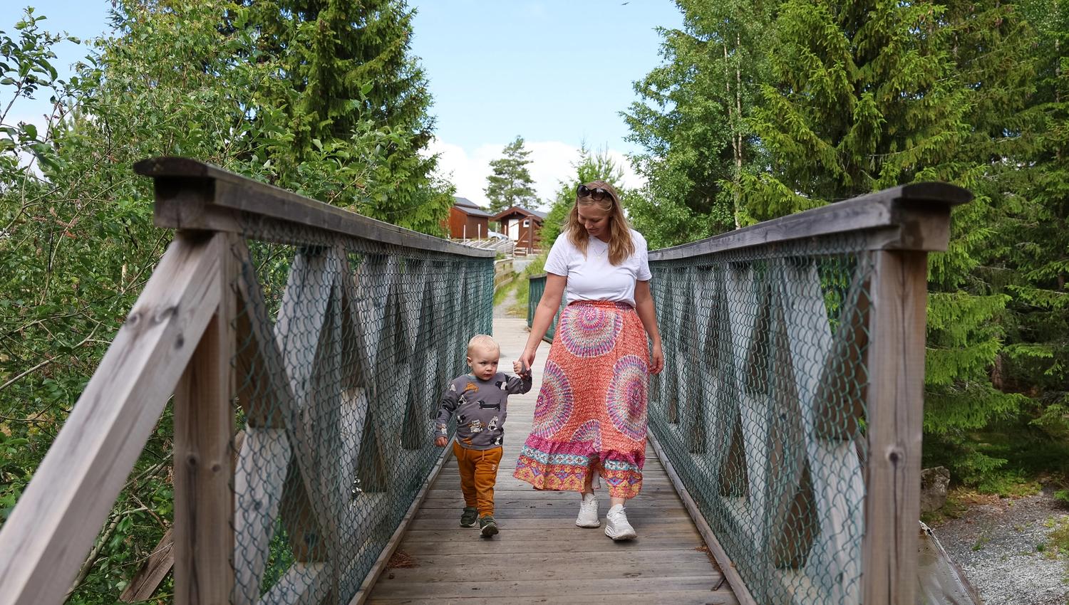 Child and an adult walking across a wooden bridge surrounded by green forest in the nature park.
