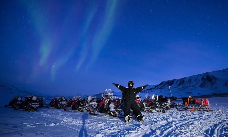 A person is sitting on a snowmobiles, with many other snowmobiles around. The northern lights are dancing on the sky.