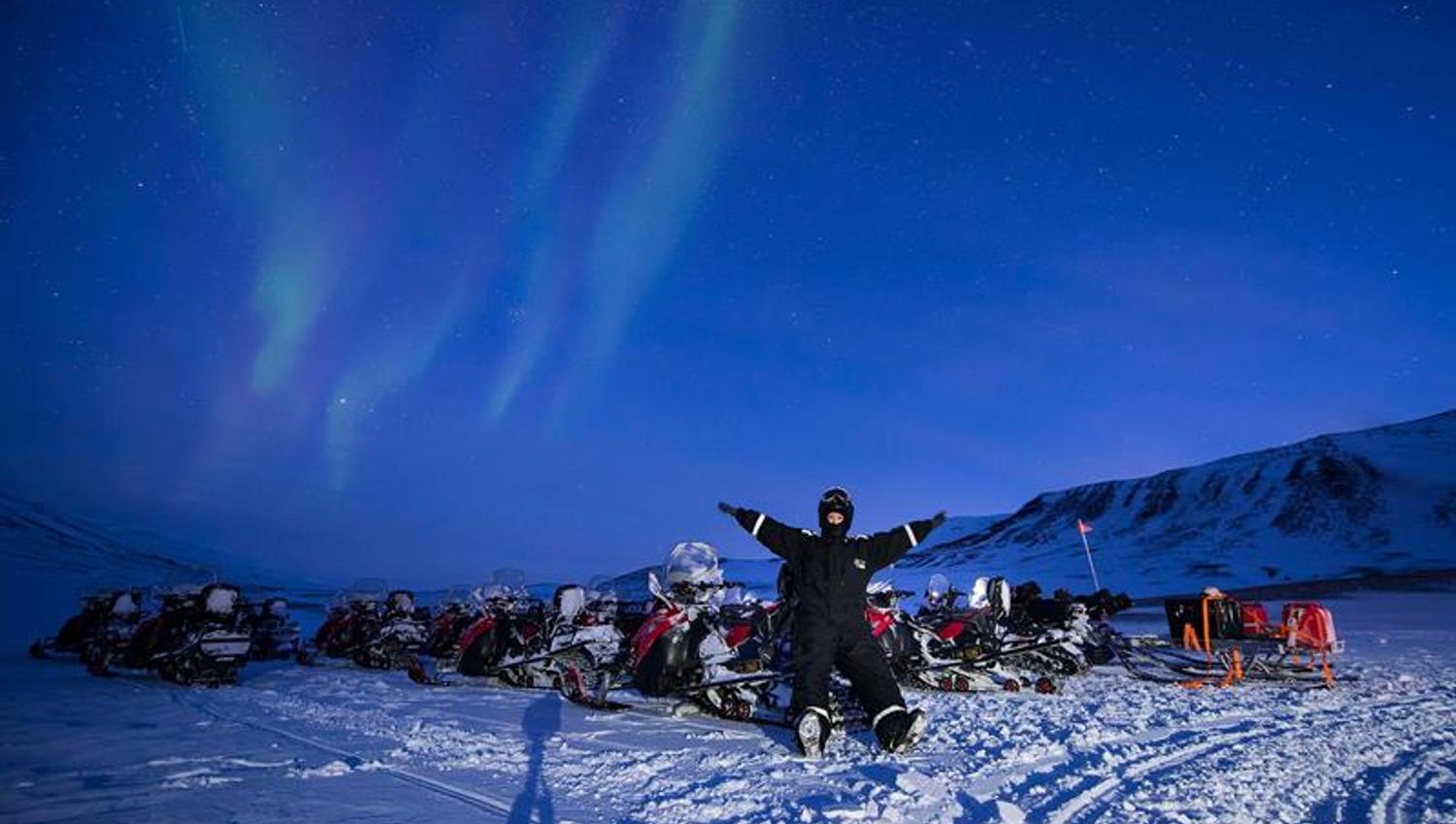 A person is sitting on a snowmobiles, with many other snowmobiles around. The northern lights are dancing on the sky.
