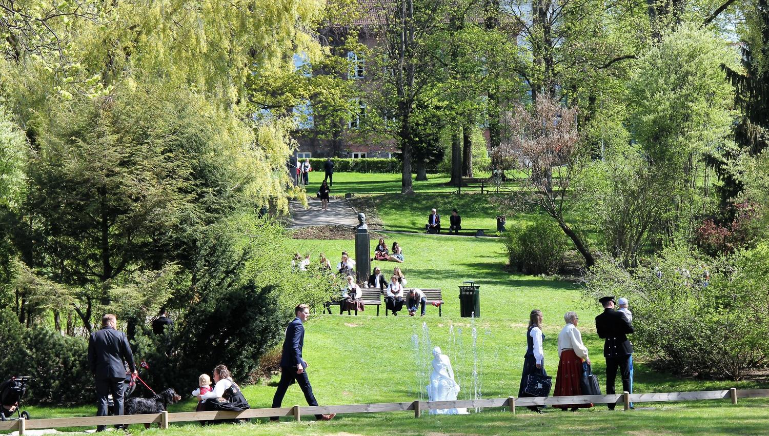 Grønn park med trær, gress og benker, der flere mennesker går og sitter mens en liten fontene spruter vann..