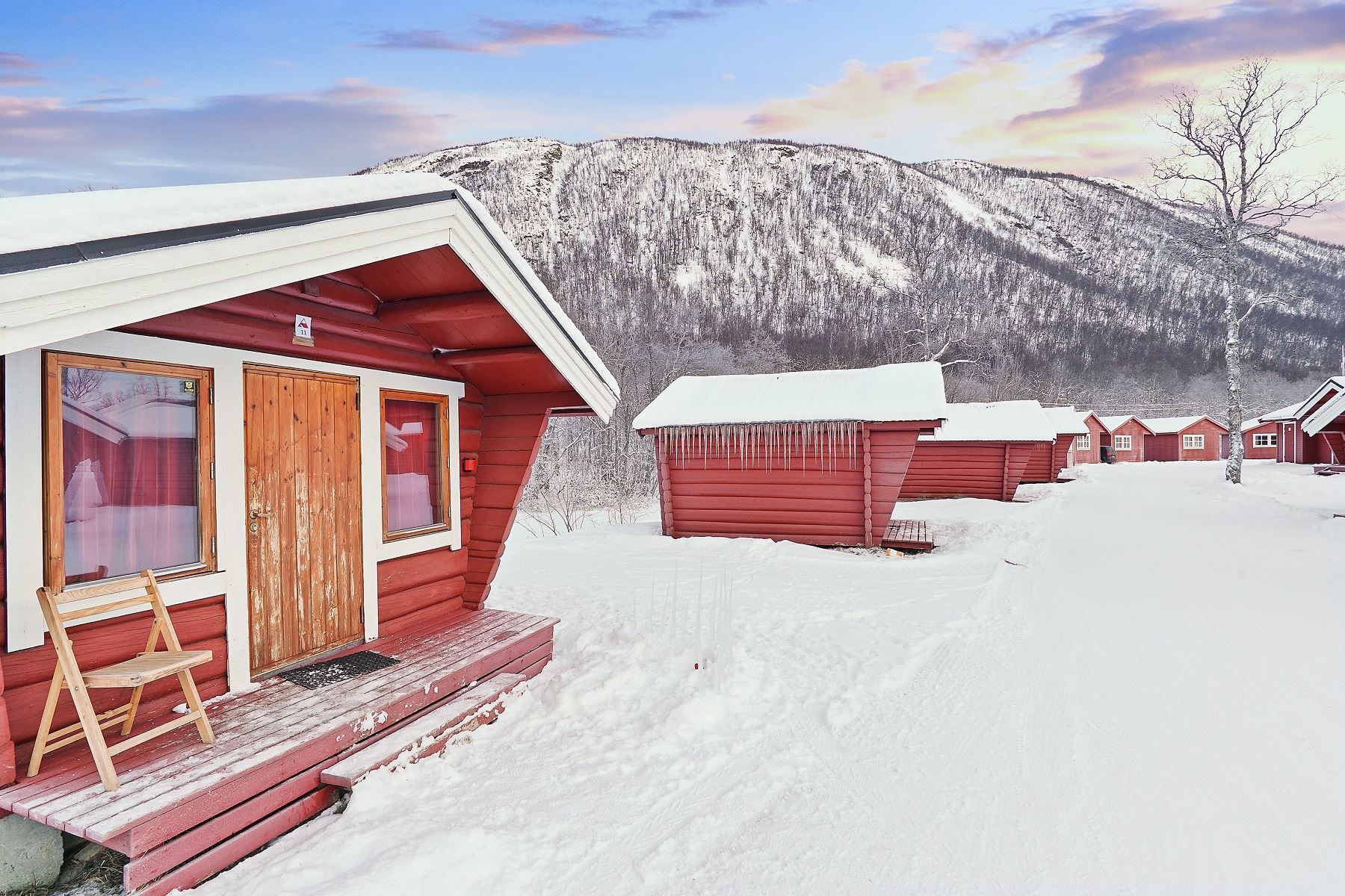 Red wooden cabins in a snowy landscape with mountains and colourful sky in the background