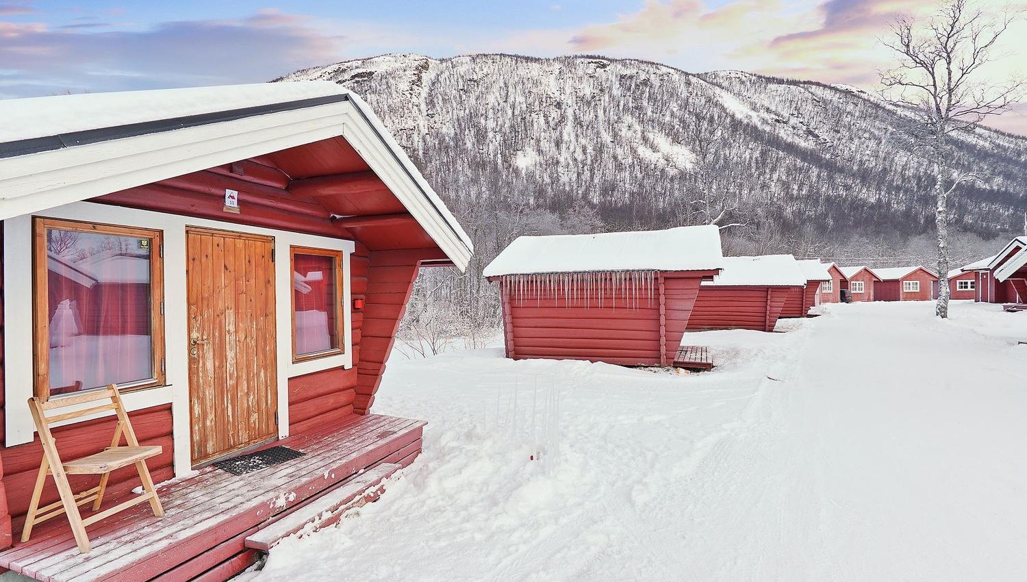 Red wooden cabins in a snowy landscape with mountains and colourful sky in the background