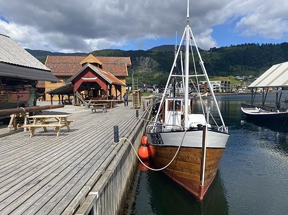 A wooden boat docked at the pier at Hardanger Maritime Center, with mountains and landscape in the background.