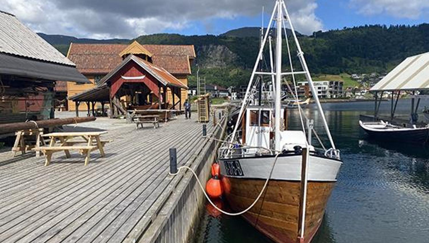 A wooden boat docked at the pier at Hardanger Maritime Center, with mountains and landscape in the background.