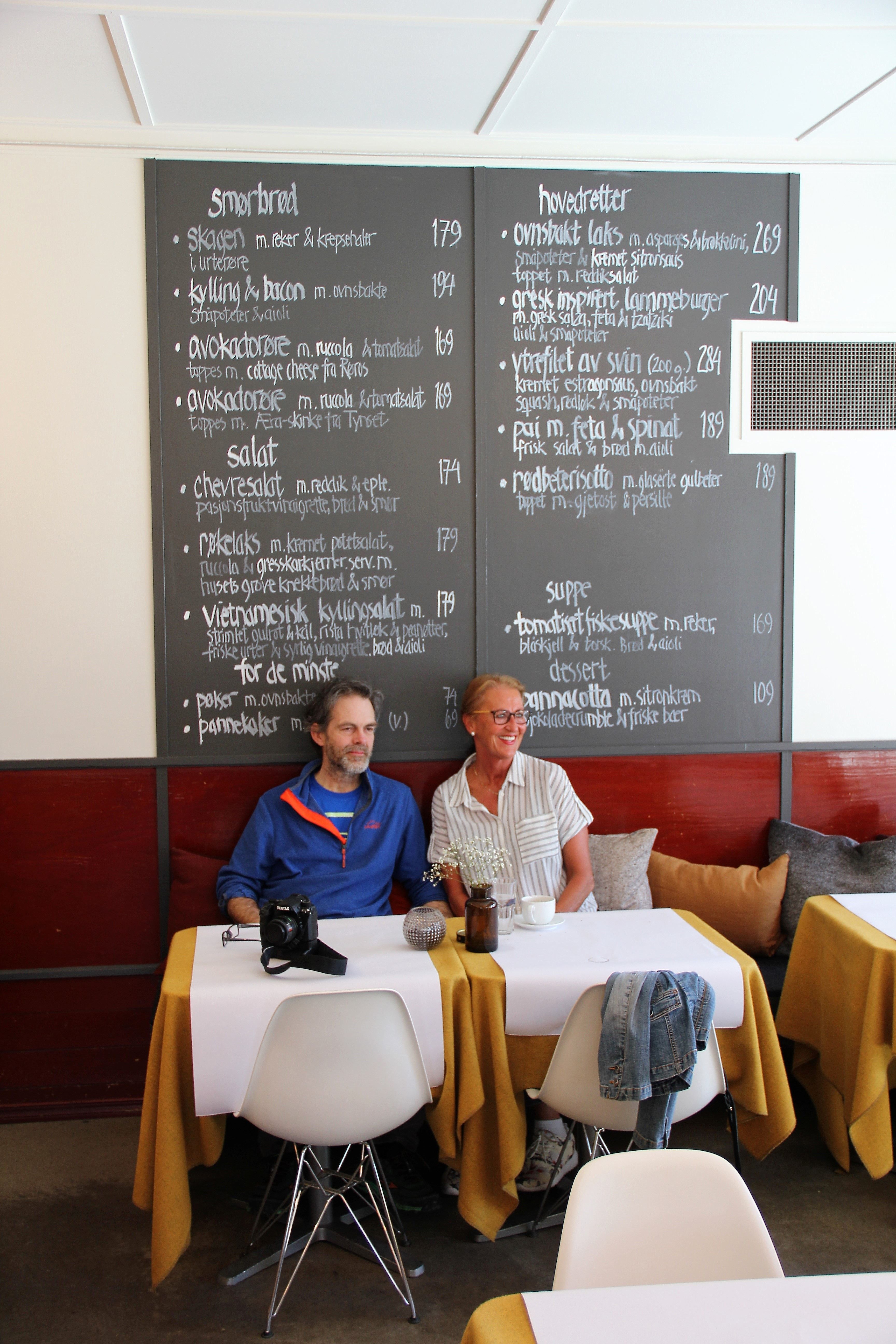 To personer sitter ved et bord i en kafé, foran en stor tavlemeny på veggen, med kopper og kamera på bordet..