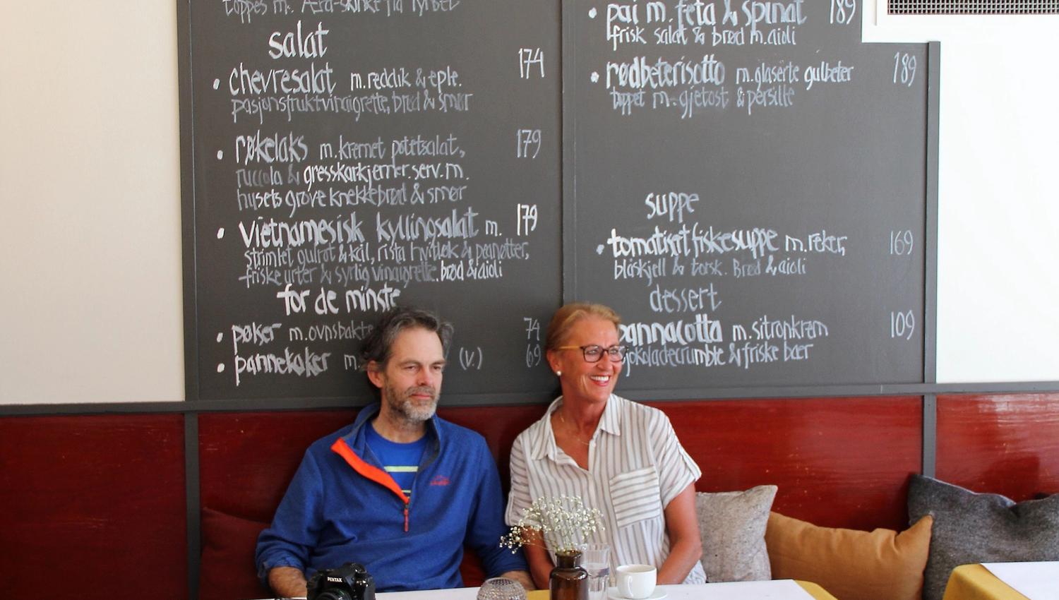 To personer sitter ved et bord i en kafé, foran en stor tavlemeny på veggen, med kopper og kamera på bordet..