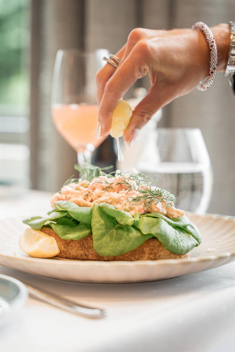 A hand squeezes lemon over an open prawn sandwich topped with fresh herbs and salad leaves.
