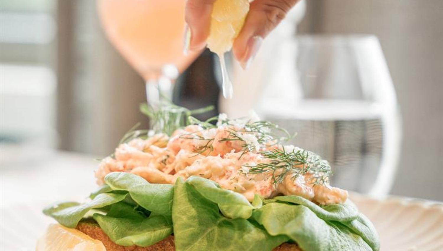 A hand squeezes lemon over an open prawn sandwich topped with fresh herbs and salad leaves.
