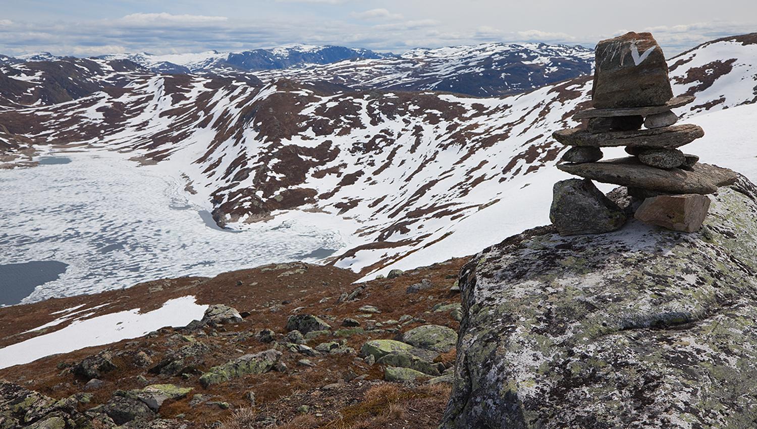 A cairn of stones with a white V markes the path to Horntind. It's early in the season, and the mountain landscape beyond is still largely snow-covere