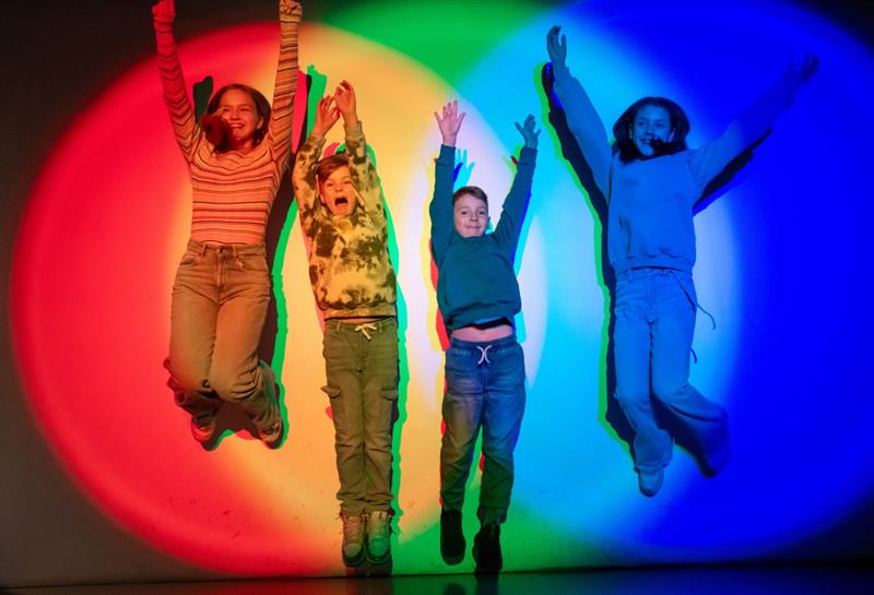 Fire barn hopper foran en vegg opplyst i sterke farger som skaper fargerike skygger bak dem