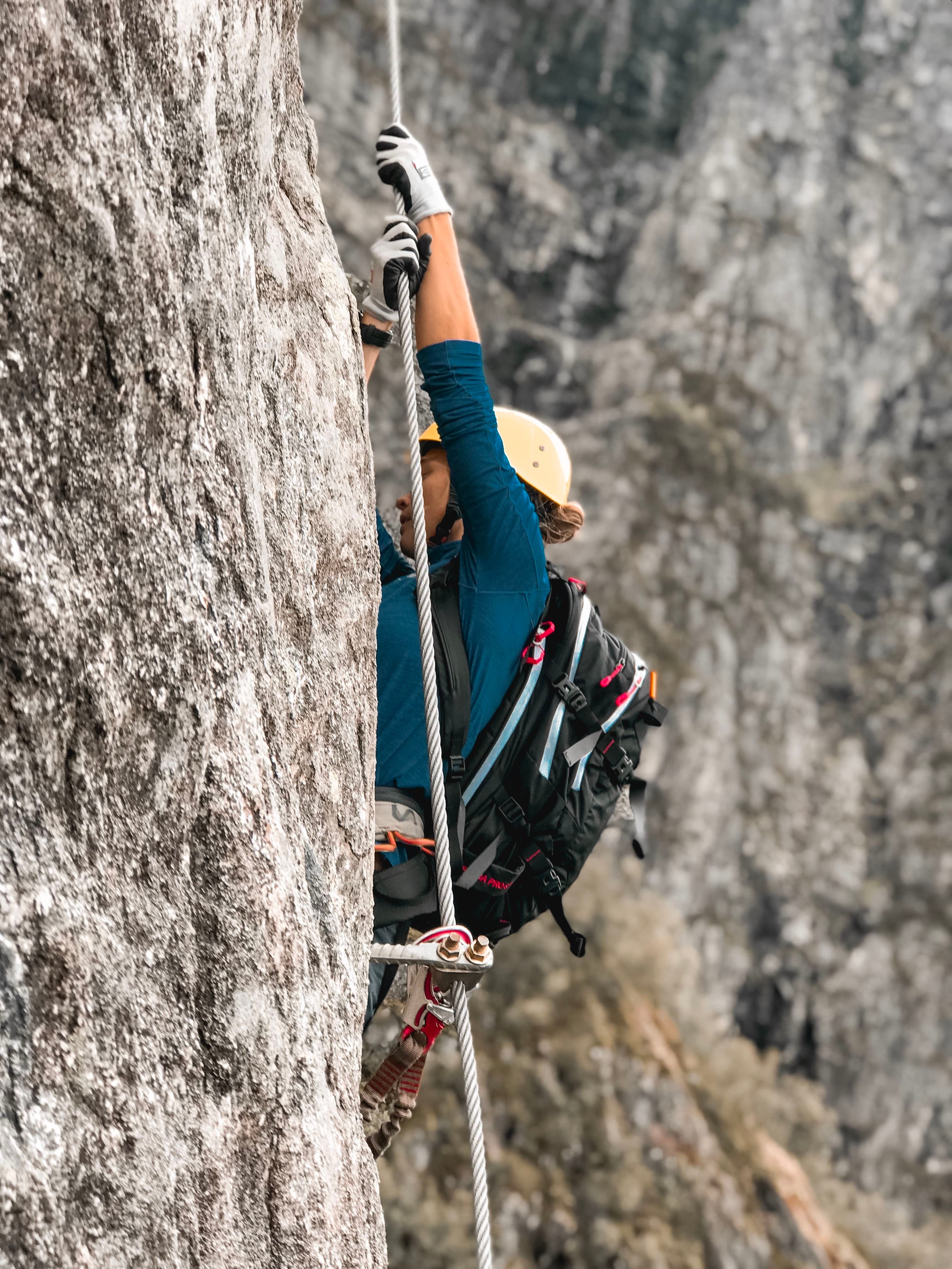 Ein fjellklatrar med hjelm og ryggsekk klatrar opp ei bratt steinvegg, sikra i tau med karabinar..