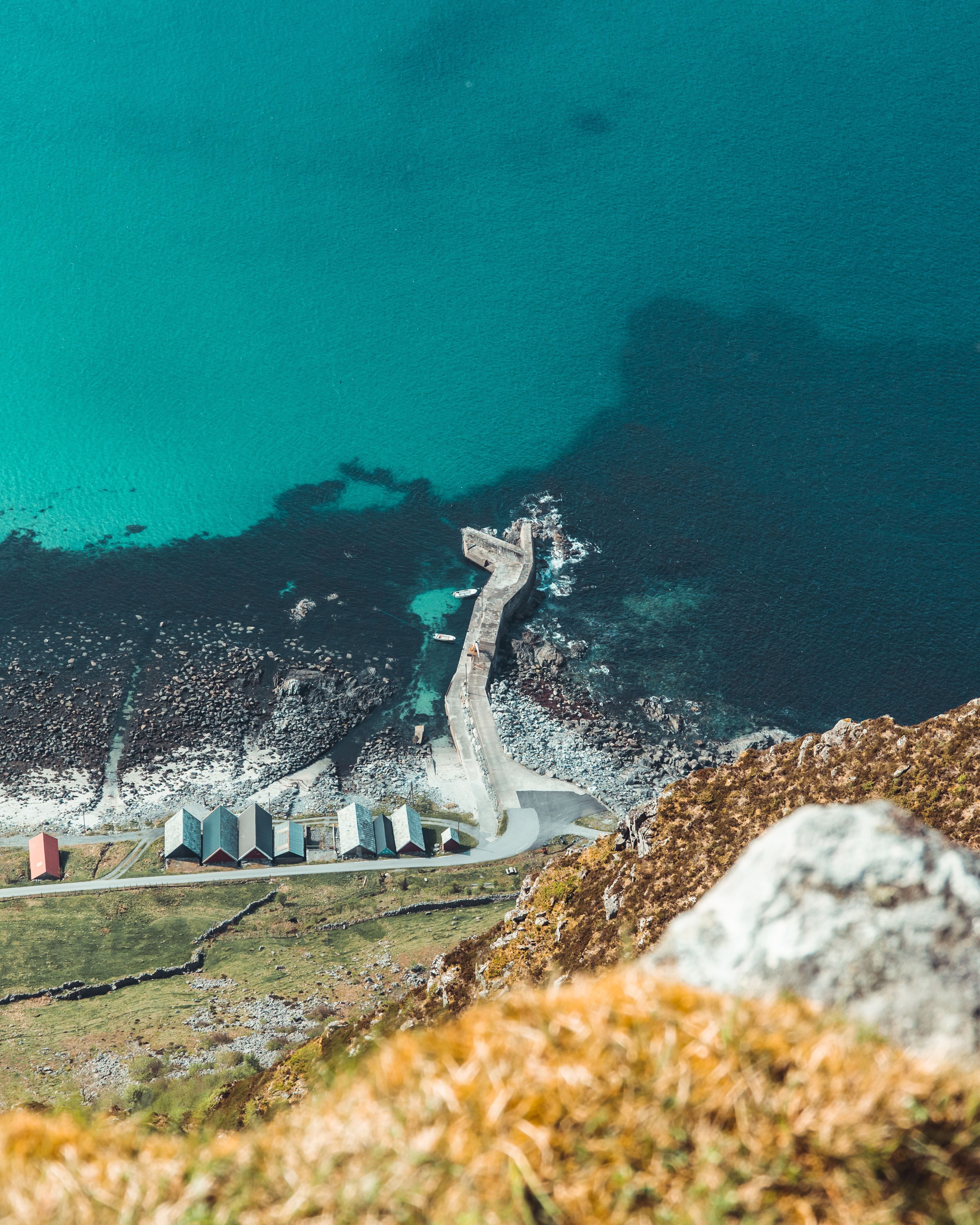 High-angle view of a rocky coastline with a narrow pier extending into clear turquoise water and small huts below..