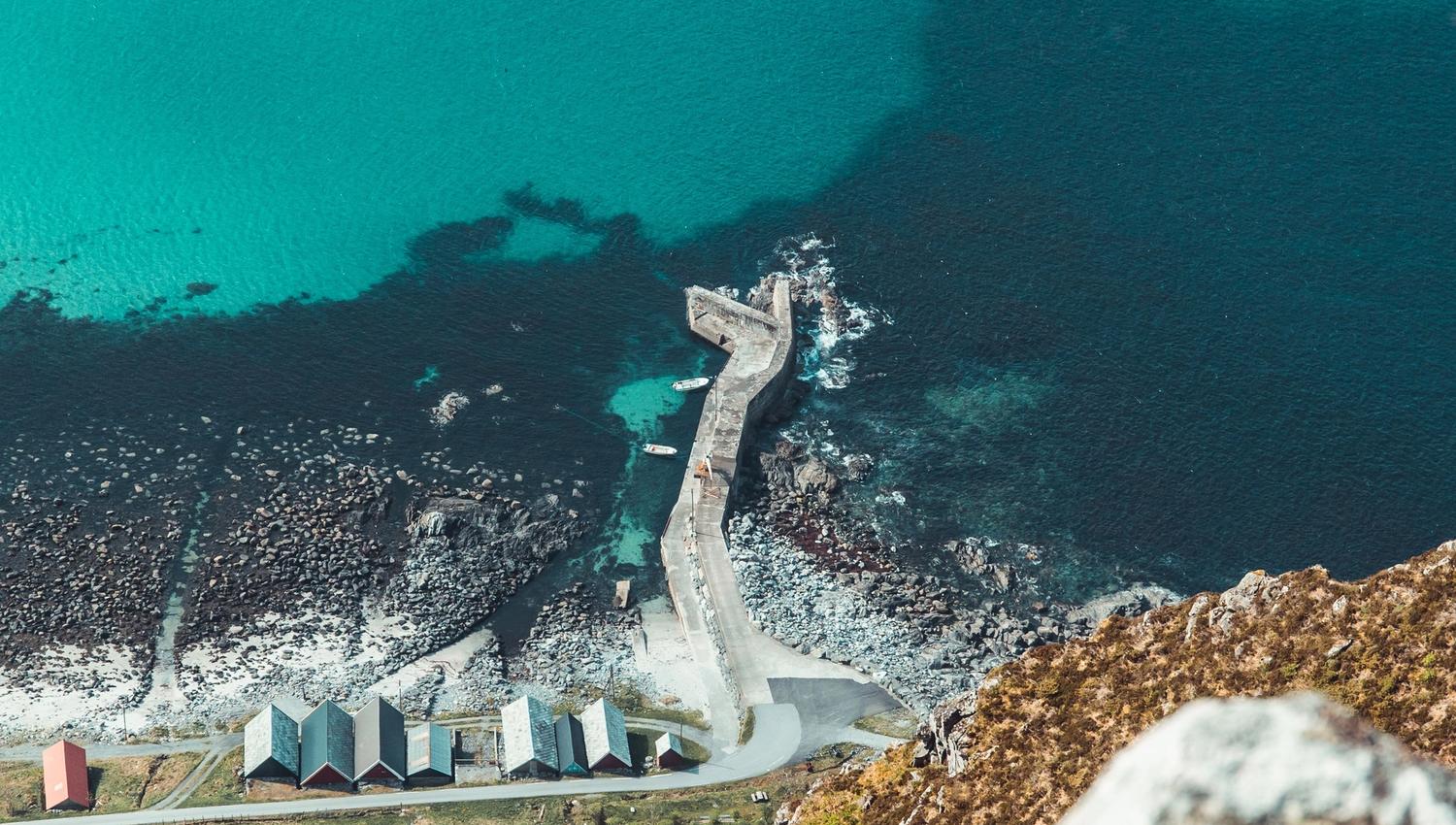 High-angle view of a rocky coastline with a narrow pier extending into clear turquoise water and small huts below..