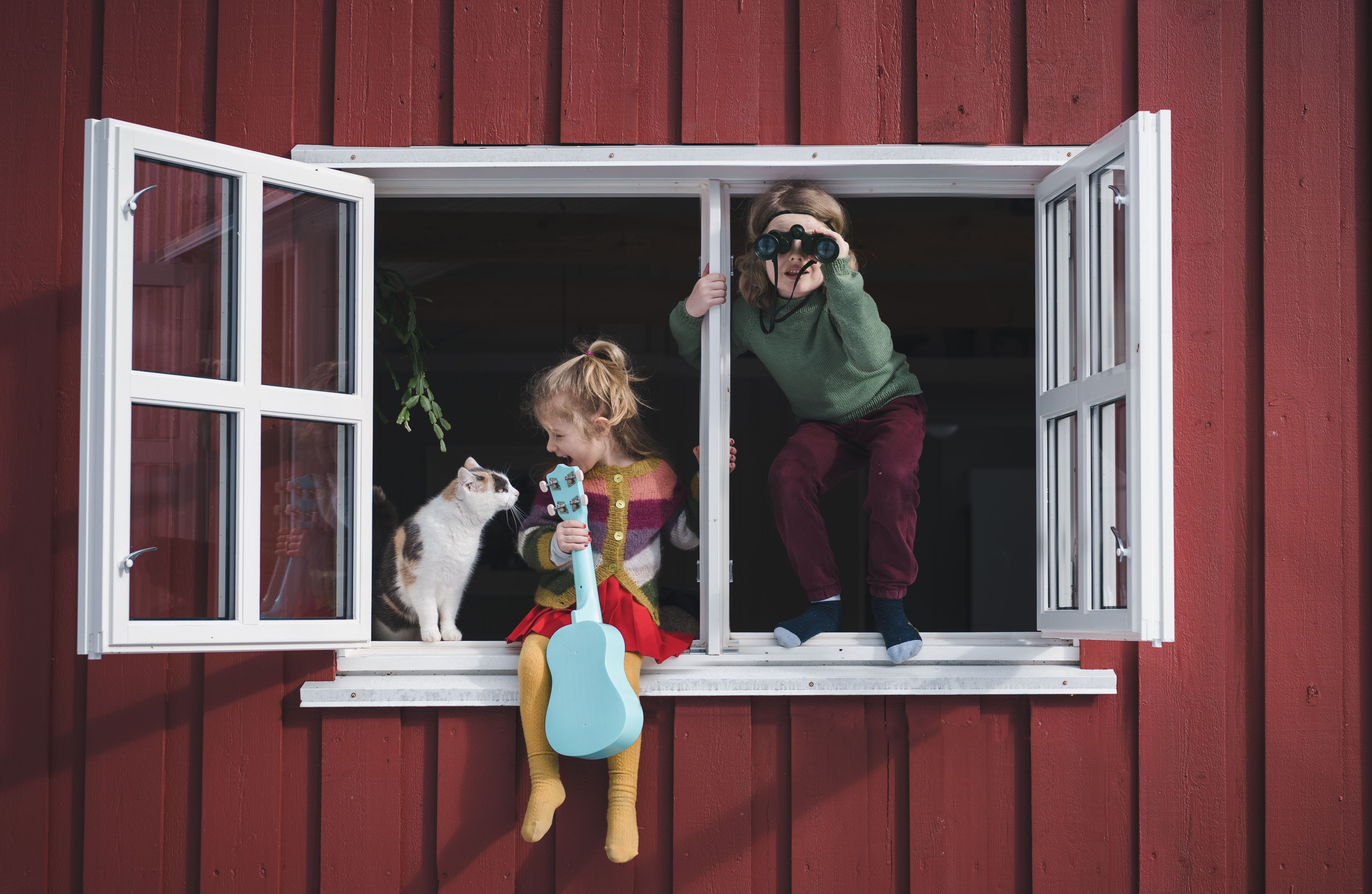 To barn sitter i vinduskarmen, den ene holder en gitar og klapper en katt, mens den andre bruker en kikkert til å se ut.